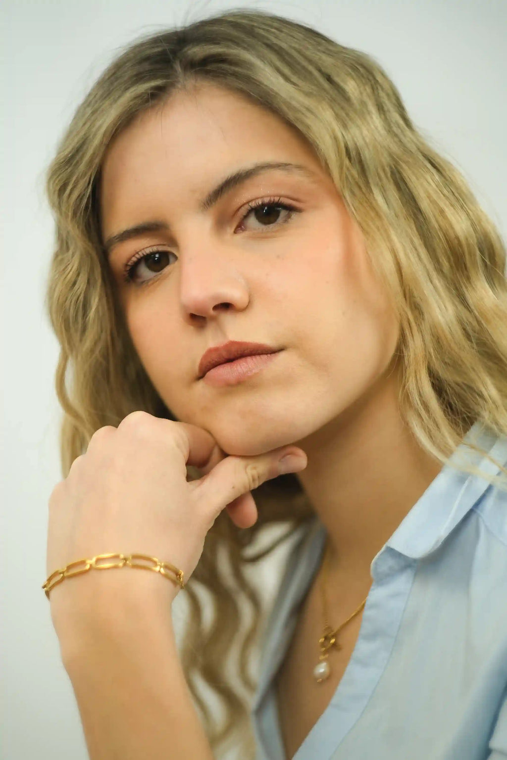 Woman with blonde hair wearing a light blue shirt and gold jewelry against a white background