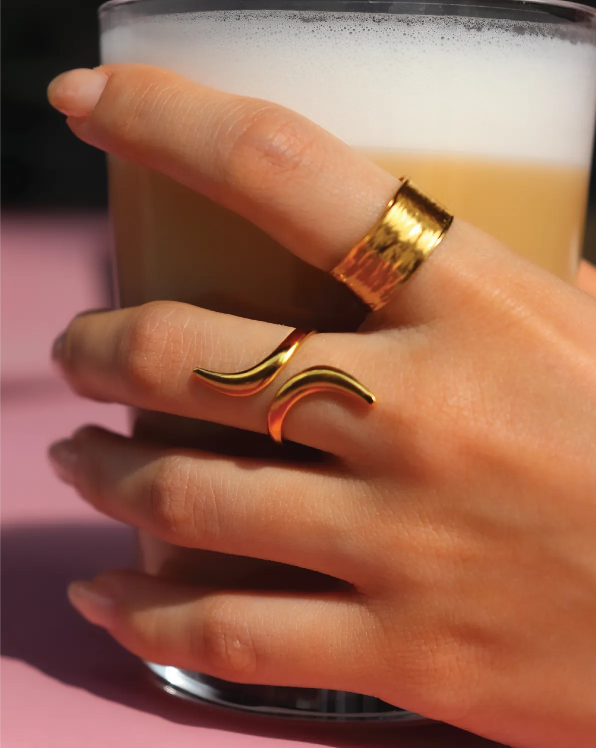 Hand wearing a gold ring holding a glass of beer on a pink surface